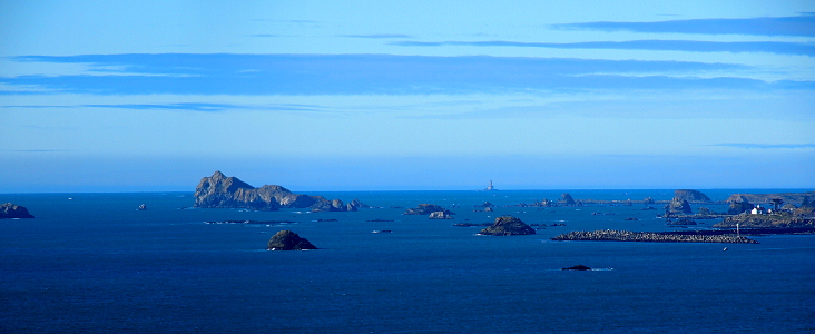 [Two photos stitched together to create a landscape image of the ocean with the St. George Reef lighthouse in the middle of the water in the distance and expanses of rocks jutting from the foreground water. The Battery Point lighthouse at the far right is near the beachhead which is not visible on this image.]